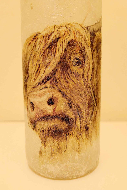 A frosted glass bottle featuring a detailed decoupage illustration of a Highland cow's face on the front, set against a plain beige background.