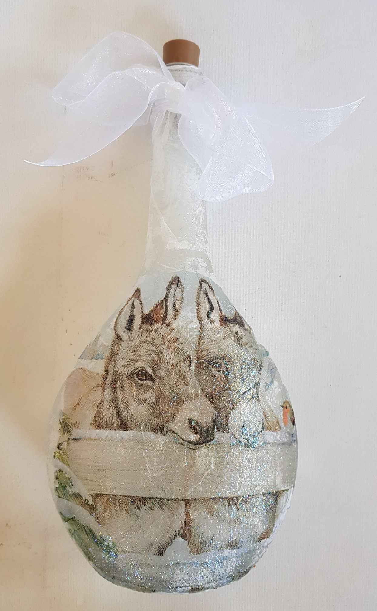 A decorative teardrop-shaped glass bottle with a cork stopper and a white organza bow at the neck, featuring a painted illustration of two donkeys behind a fence on the front.