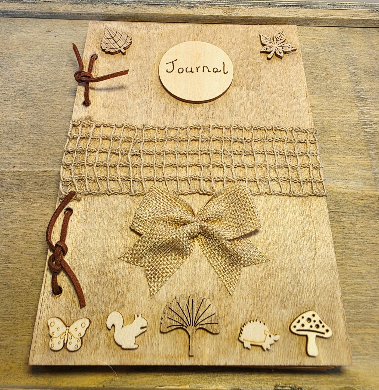 wooden journal with rustic embellishments on the cover such as leaves and woodland creatures. Burlap decoration across the middle of the journal. Resting in a wooden tray with a pen on top