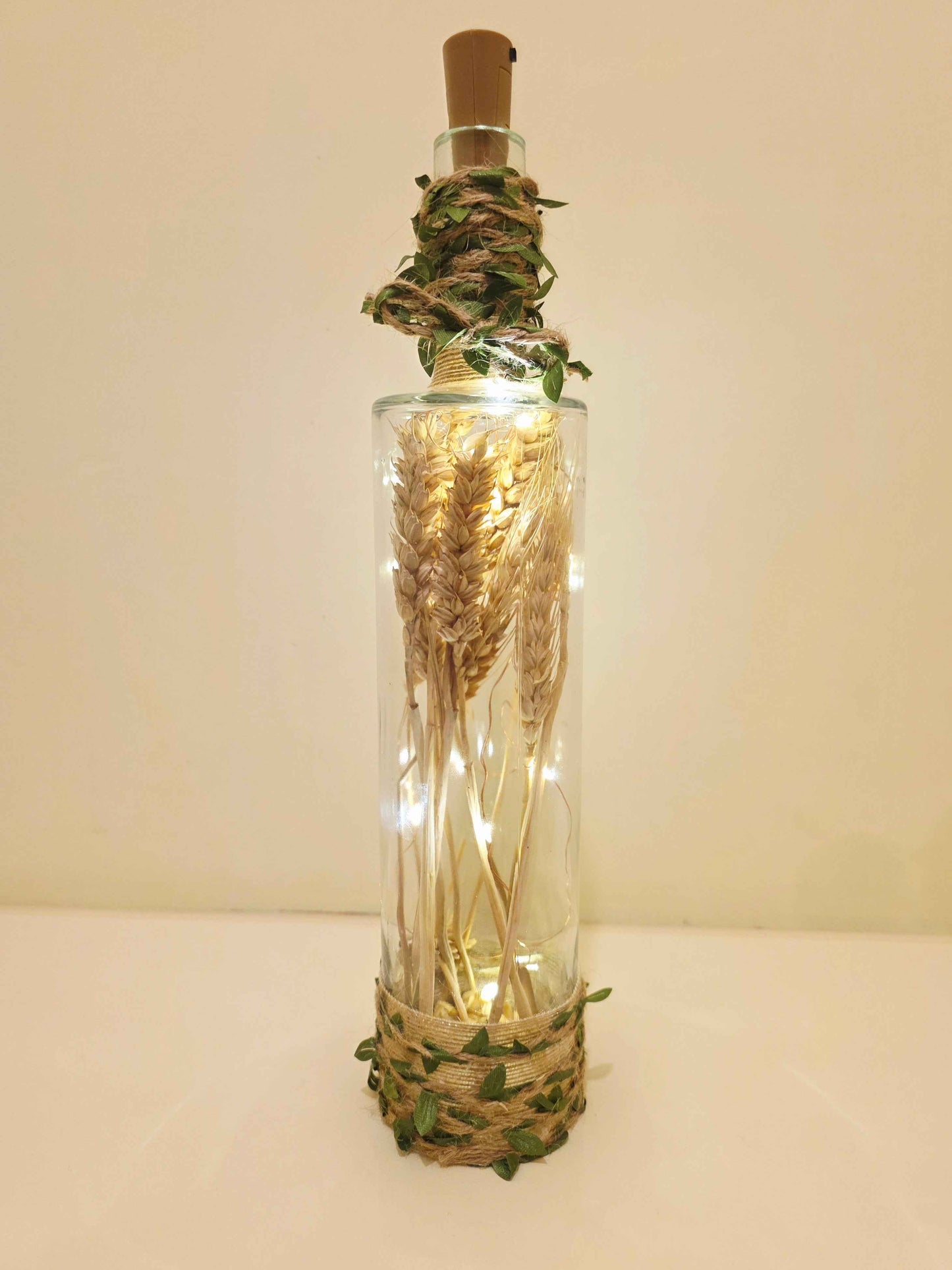 A clear glass bottle with a cork stopper, decorated with dried wheat stalks, green leafy vines, and warm white fairy lights inside, set against a plain beige background.