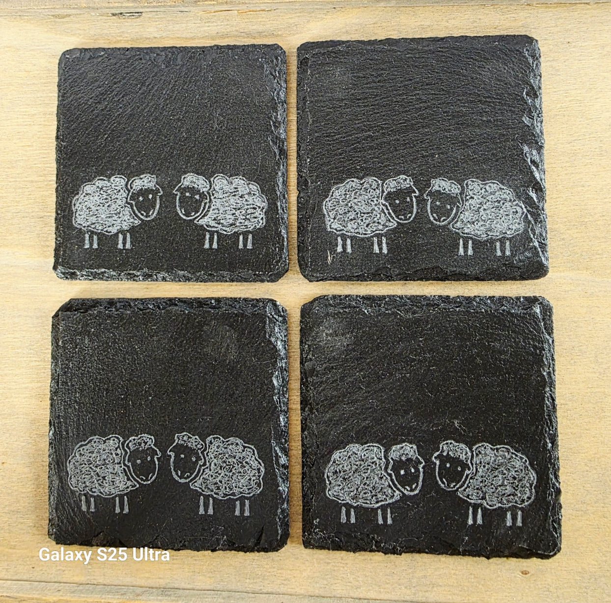 Four black slate coasters with white sheep illustrations on a wooden surface.