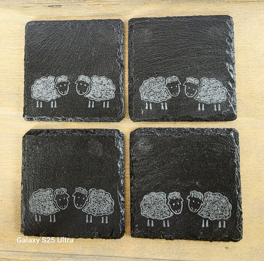 Four black slate coasters with white sheep illustrations on a wooden surface.