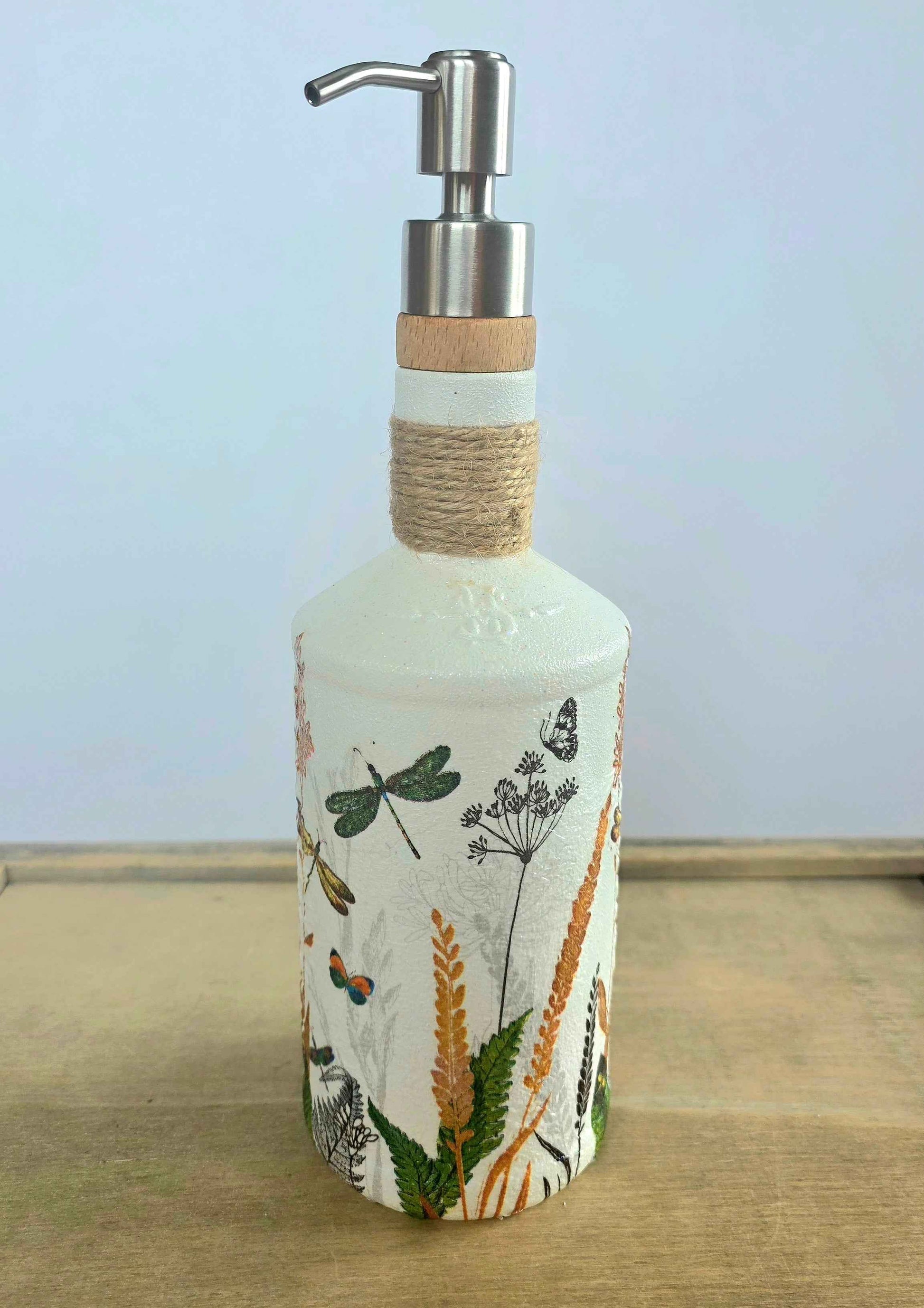 A decorative soap dispenser bottle with a white base decorated with botanical and insect illustrations, featuring a twine-wrapped neck and a stainless steel pump dispenser.
