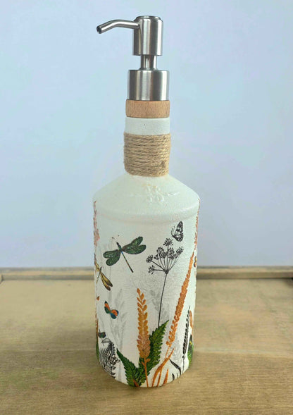 A decorative soap dispenser bottle with a white base decorated with botanical and insect illustrations, featuring a twine-wrapped neck and a stainless steel pump dispenser.