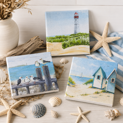 Three square canvas coastals with beach-themed paintings are arranged on a light wood surface with starfish, shells, driftwood, and a striped cloth. The canvases depict a lighthouse, seagulls on a dock, and small beach huts.