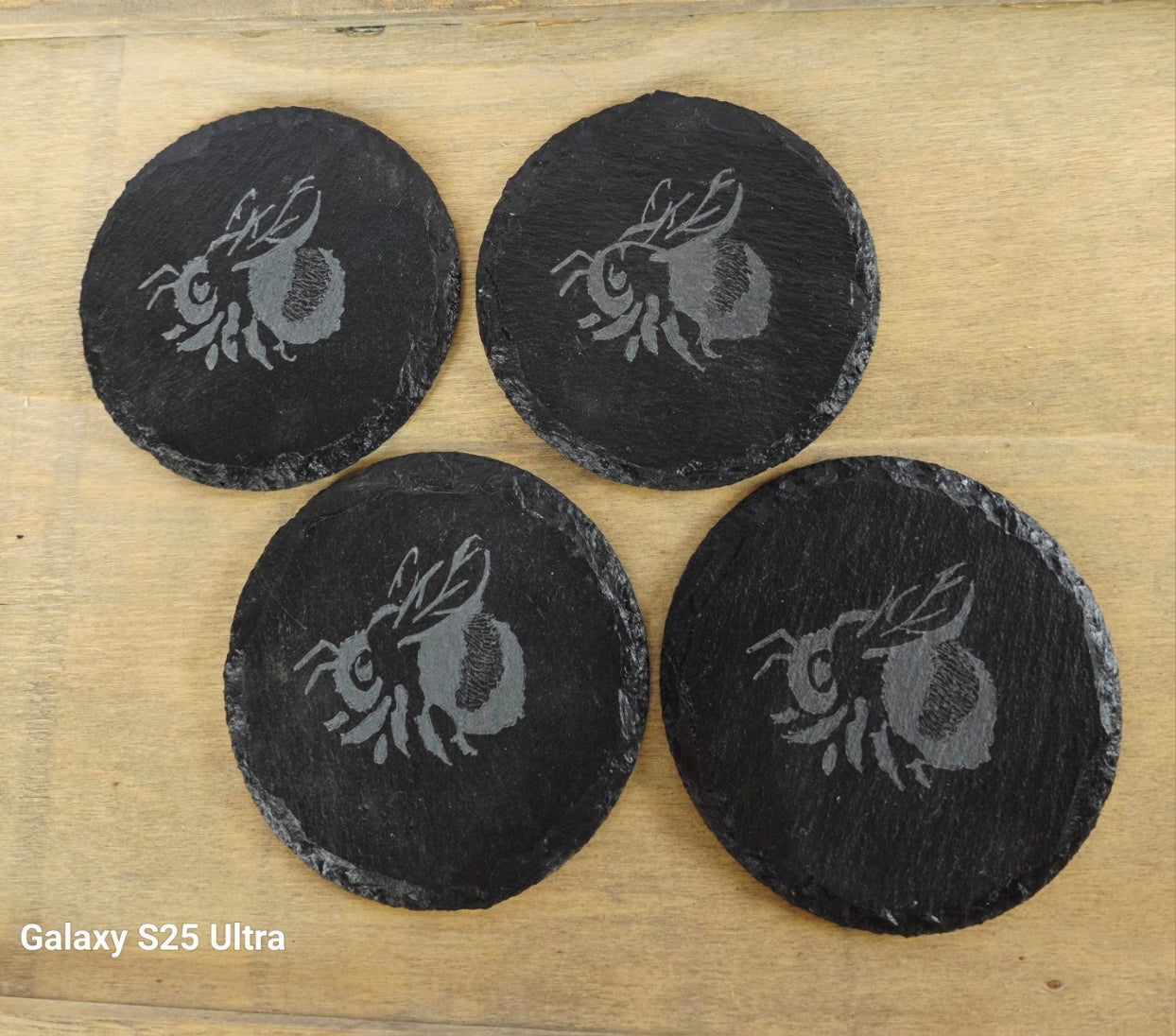 Four black coasters with a engraved bee design on a wooden surface