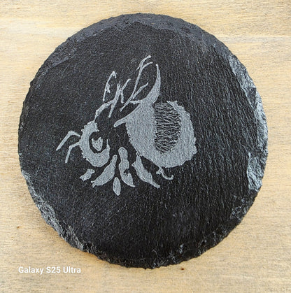 Round black slate coaster with engraved bee design on a wooden surface