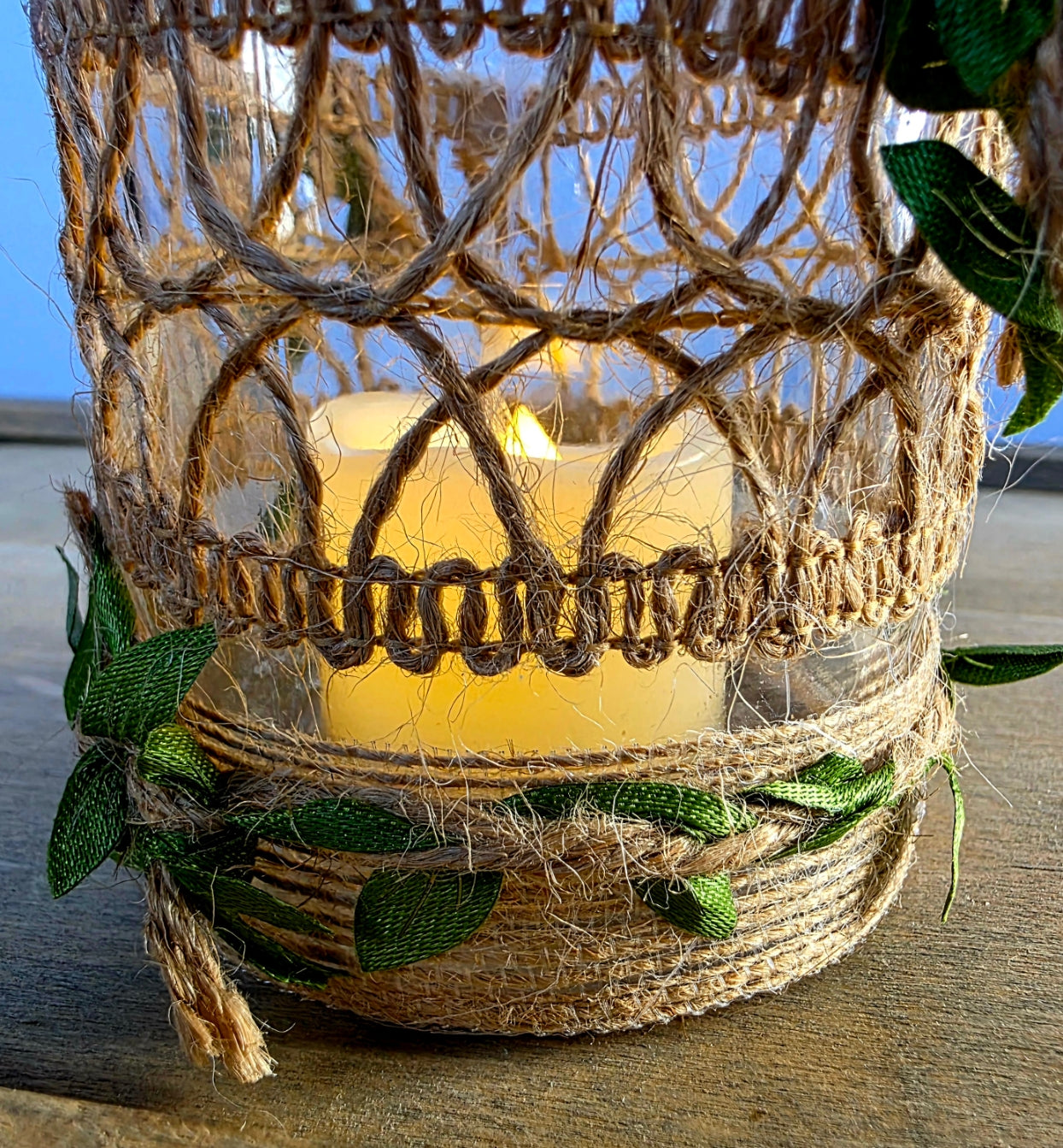 Decorative lantern with a flameless candle inside, decorated with burlap twine with green satin leaves, on a wooden surface.
