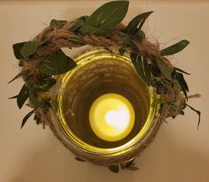 Flameless Candle in a glass jar wrapped with twine and green leaves on a neutral background