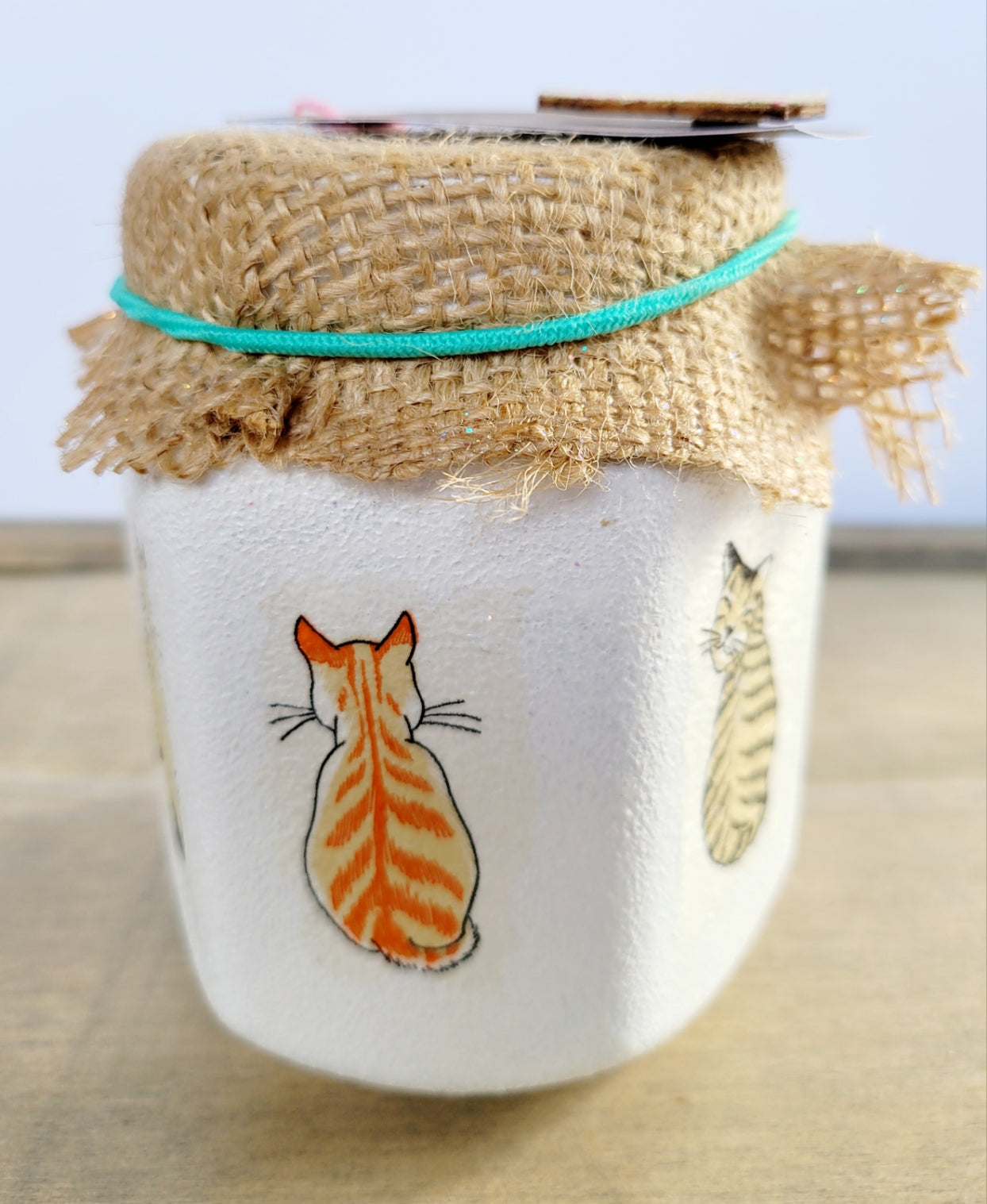 white jar with a marmalade cat and tabby cat looking over its shoulder. Brown hessian lid secured with green elastic band