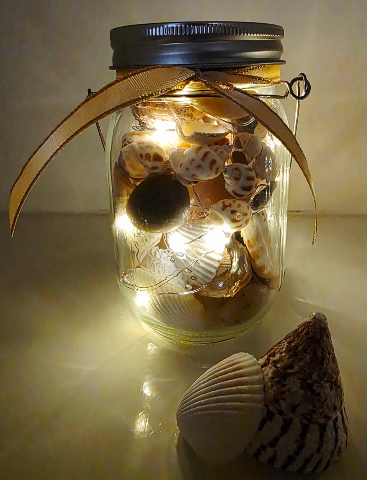 Jar filled with shells, pebbles and coloured glass stones. Lit up with lid and carry handle. gold decorative ribbon