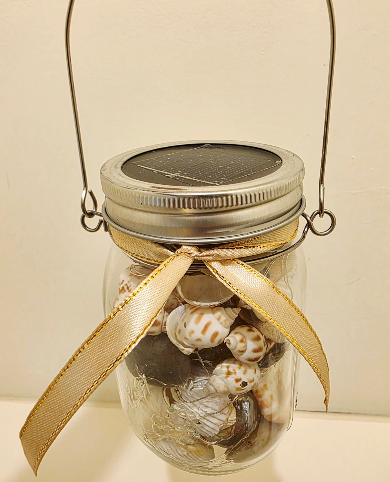 Glass jar with shells and a gold ribbon on a beige background. jar has metal carry handle