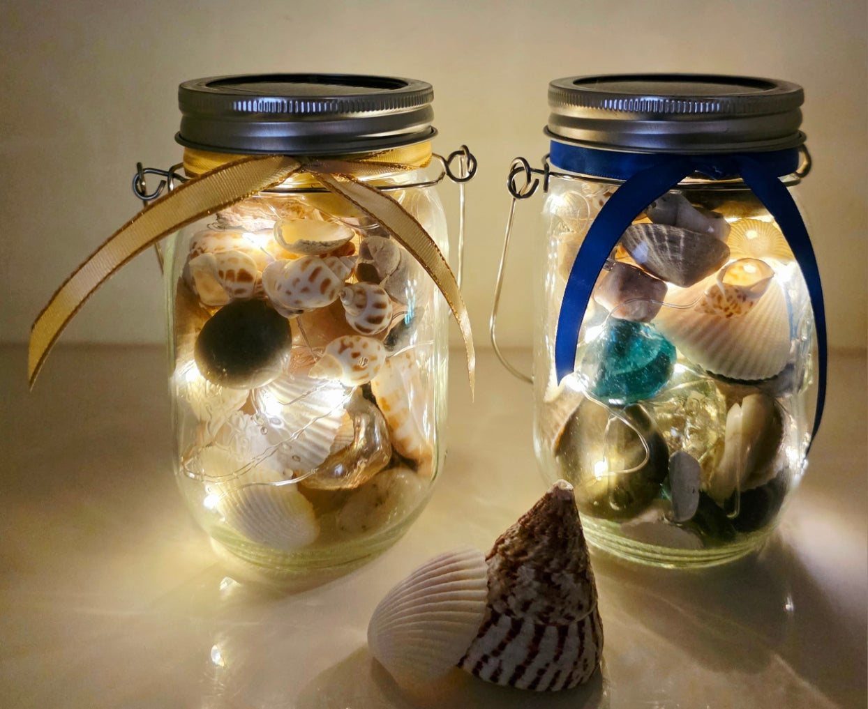 Jars filled with shells, pebbles and coloured glass stones. Lit up with lid and carry handle. Blue decorative ribbon