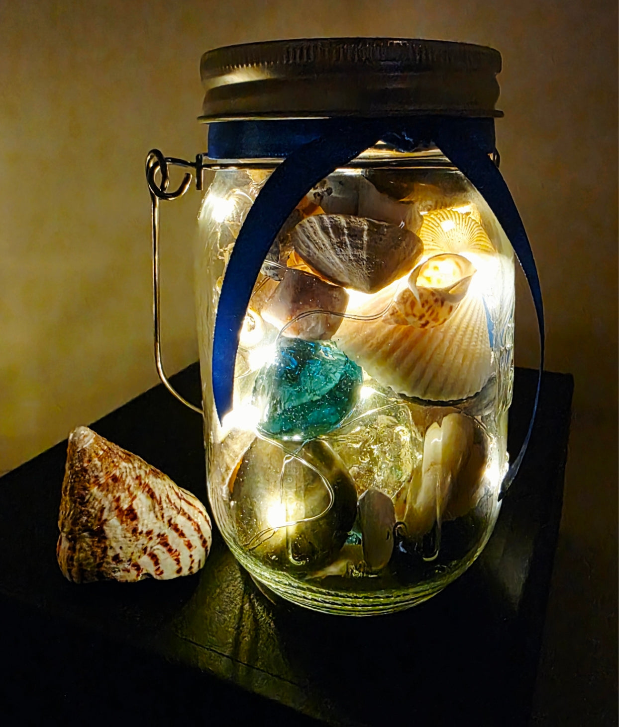 Jar filled with shells, pebbles and coloured glass stones. Lit up with lid and carry handle. Blue decorative ribbon