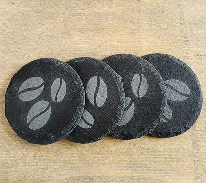 Four black slate coasters with coffee bean design on a wooden surface