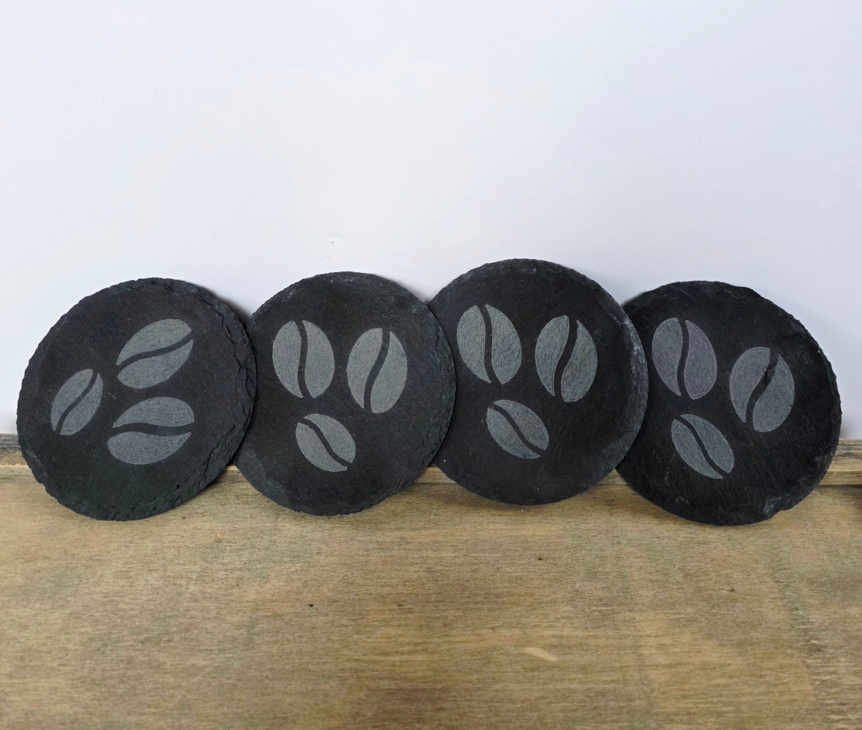 Four black, round slate coasters with coffee bean design on a wooden surface
