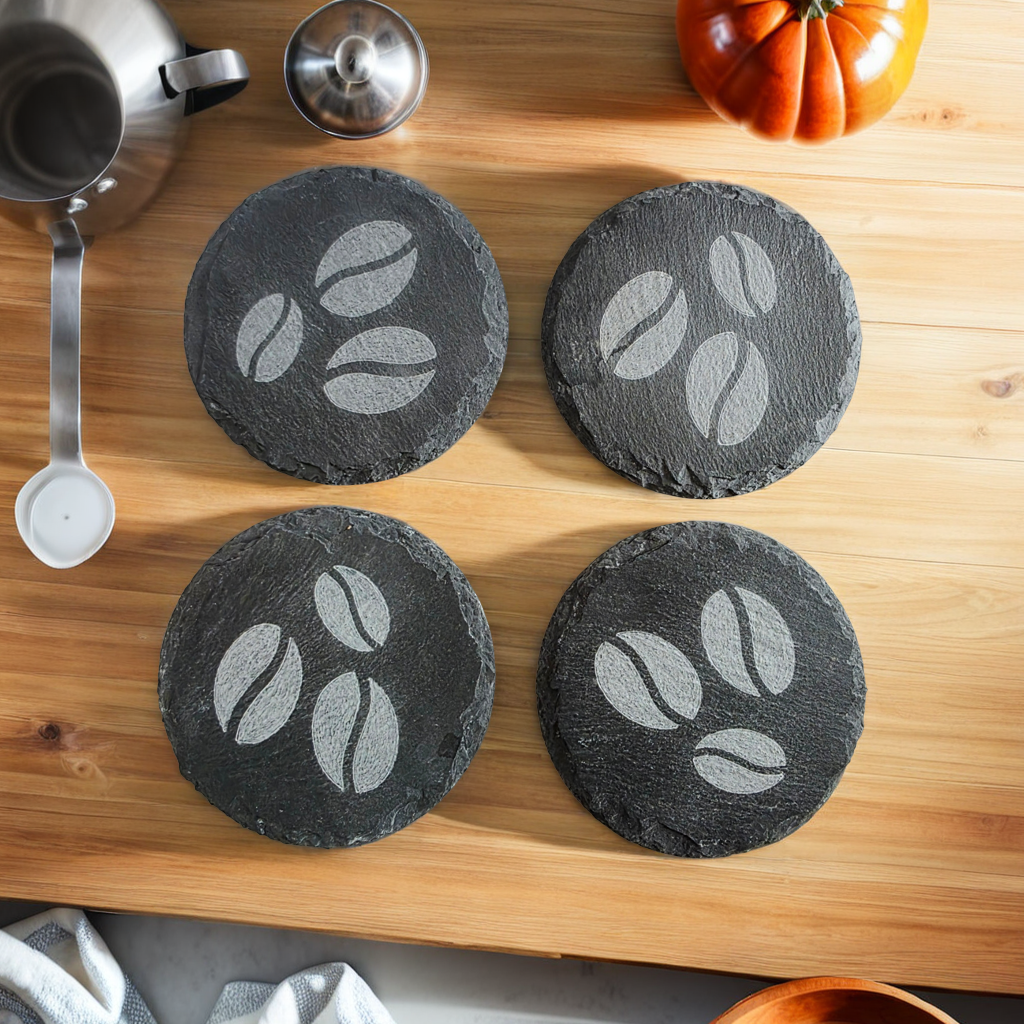 Four round, black slate coasters with coffee bean design on a wooden surface.
