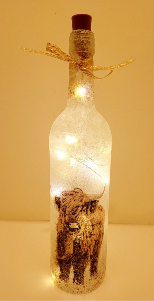 Lit up standard shape wine bottle with highland cow detailing on the front near the bottom. Gold ribbon tied around the neck of the bottle. Cork LED lights