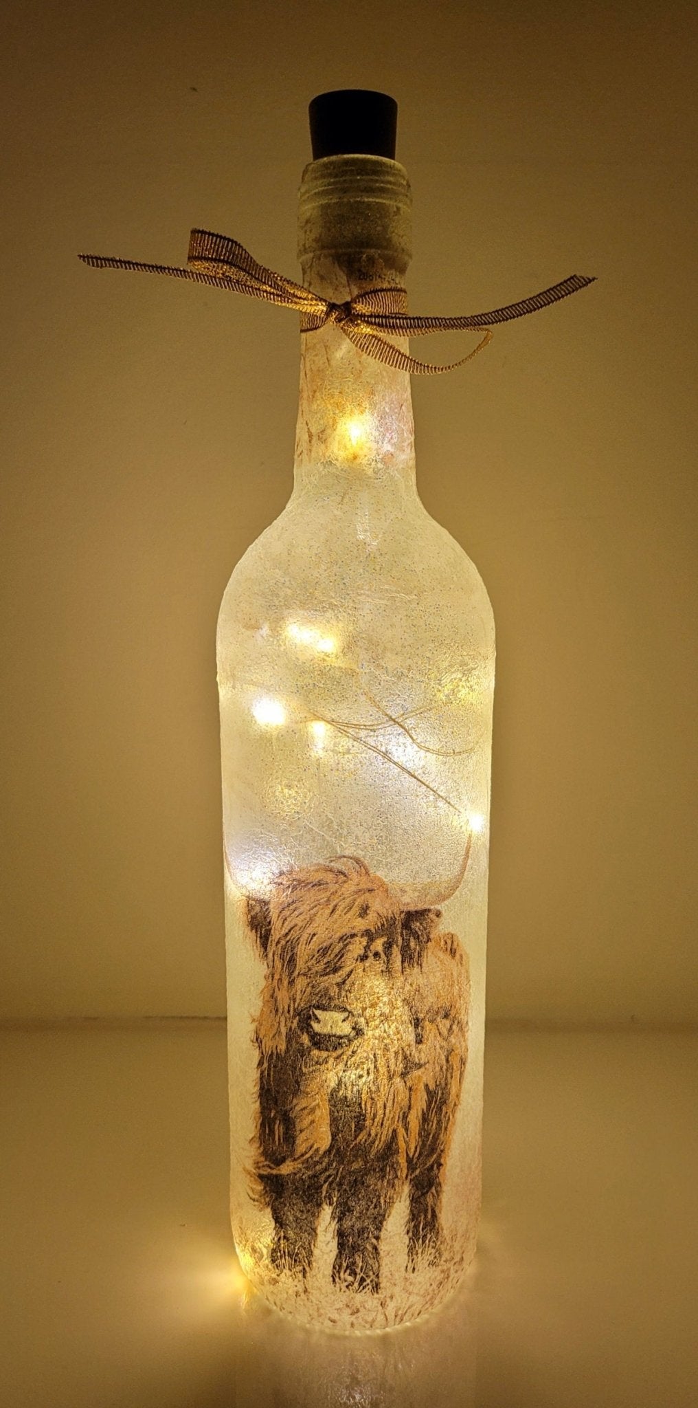Lit up standard shape wine bottle with highland cow detailing on the front near the bottom. Gold ribbon tied around the neck of the bottle. Cork LED lights
