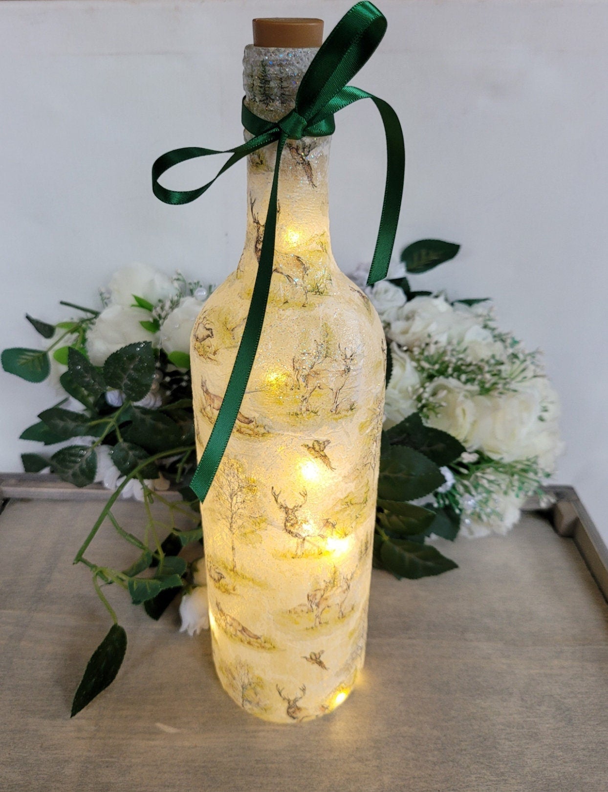 A decorative wine bottle with a woodland scene design. The bottle is illuminated with fairy lights and features a cork stopper and a dark green ribbon. The bottle's surface is textured and covered with images of deer, trees, and other forest animals.