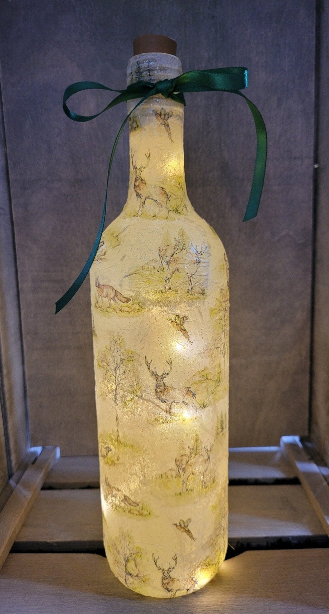 A decorative wine bottle with a woodland scene design. The bottle is illuminated with fairy lights and features a cork stopper and a dark green ribbon. The bottle's surface is textured and covered with images of deer, trees, and other forest animals.