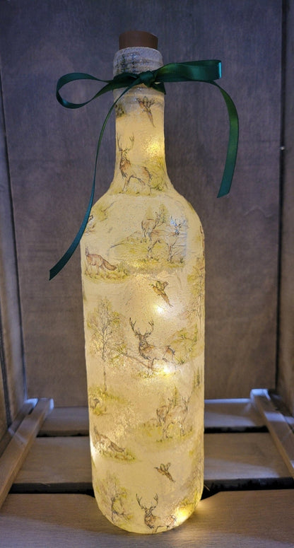 A decorative wine bottle with a woodland scene design. The bottle is illuminated with fairy lights and features a cork stopper and a dark green ribbon. The bottle's surface is textured and covered with images of deer, trees, and other forest animals.