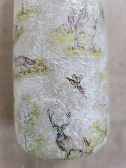 A decorative wine bottle with a woodland scene design. The bottle's surface is textured and covered with images of deer, trees, and other forest animals.