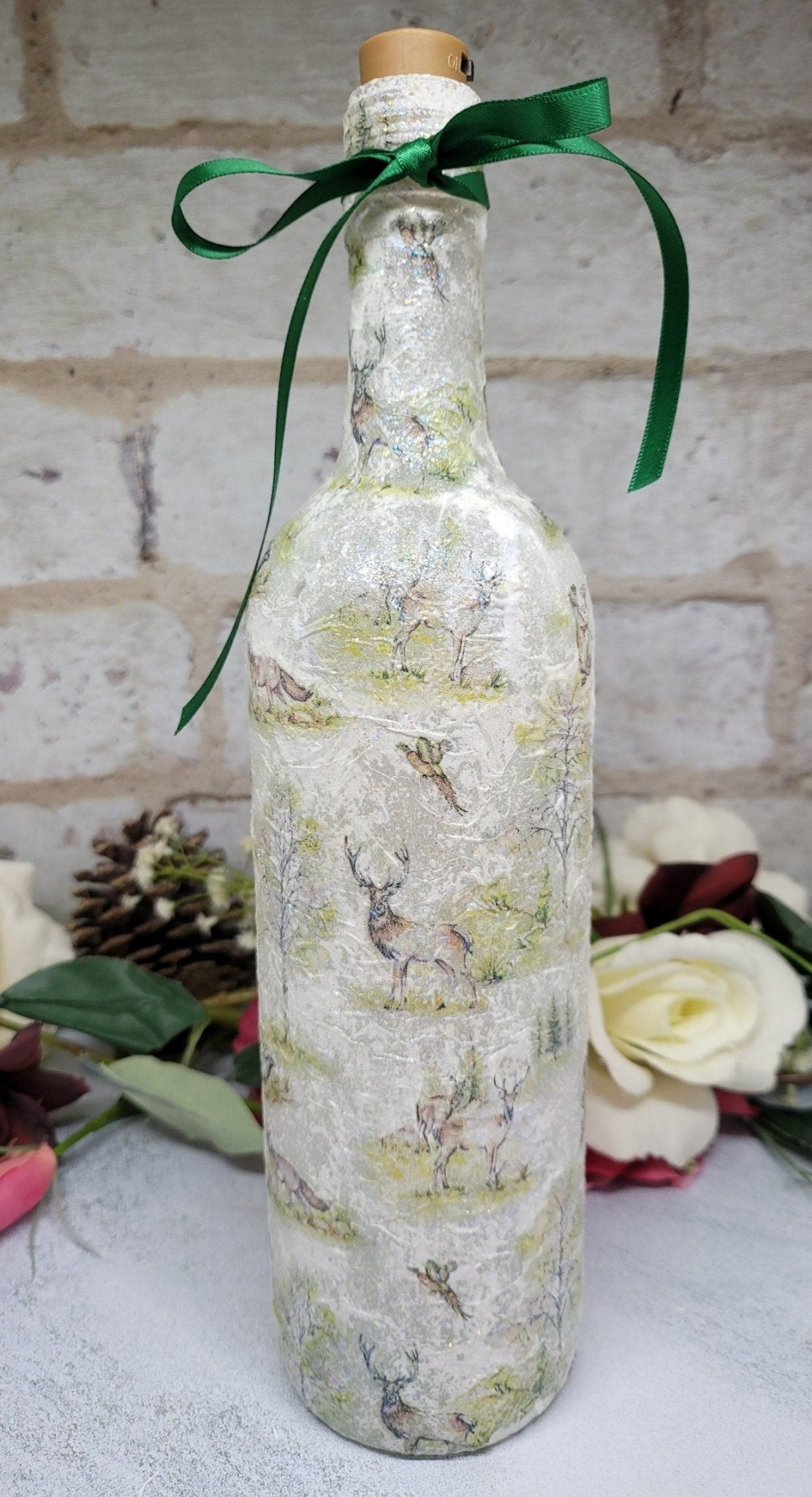 A decorative wine bottle with a woodland scene design. The bottle is illuminated with fairy lights and features a cork stopper and a dark green ribbon. The bottle's surface is textured and covered with images of deer, trees, and other forest animals.