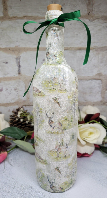 A decorative wine bottle with a woodland scene design. The bottle is illuminated with fairy lights and features a cork stopper and a dark green ribbon. The bottle's surface is textured and covered with images of deer, trees, and other forest animals.