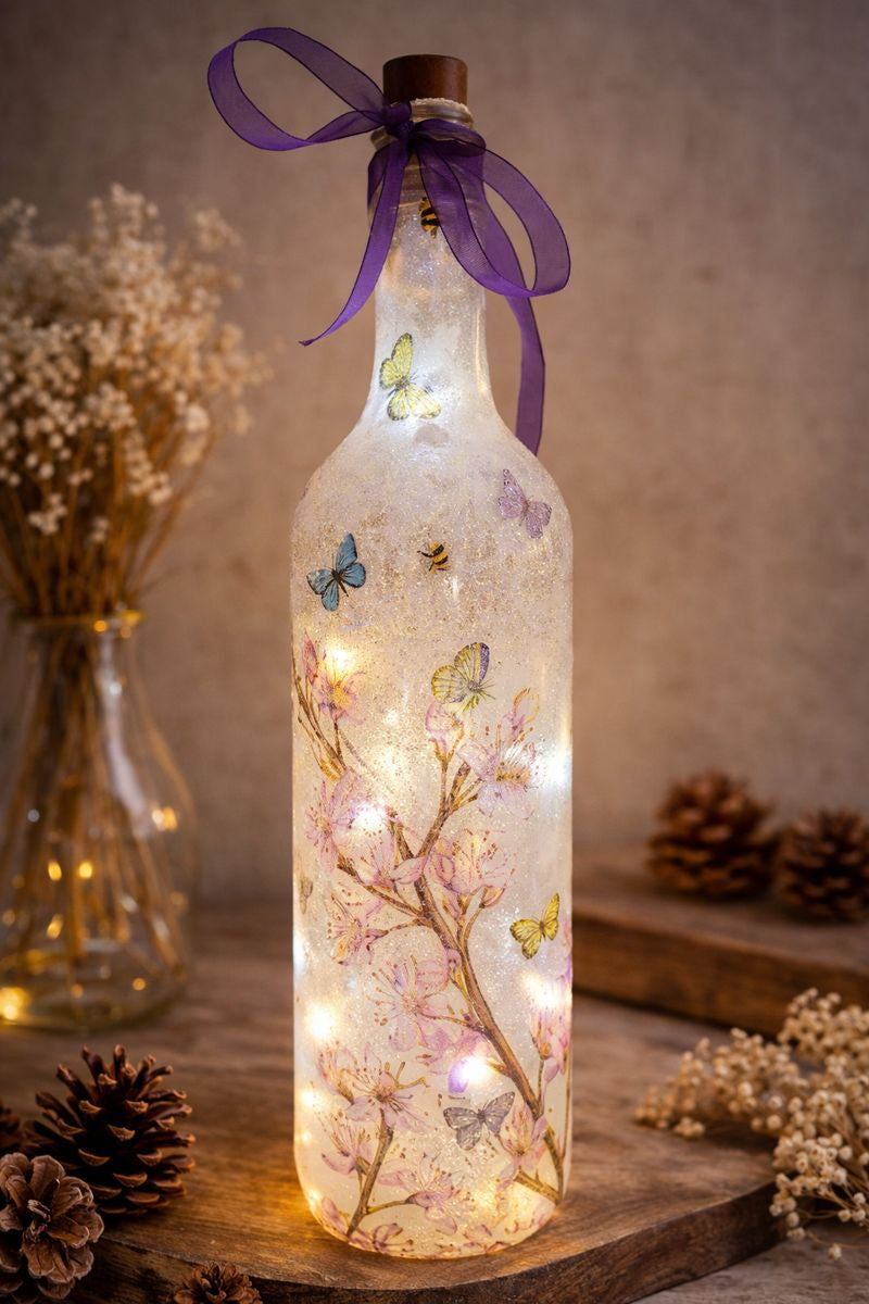 Decorative light-up bottle with floral and butterfly design, surrounded by dried plants and pinecones.