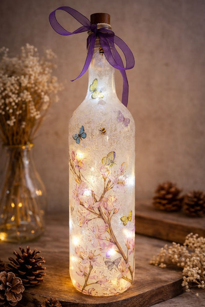 Decorative light-up bottle with floral and butterfly design, surrounded by dried plants and pinecones.