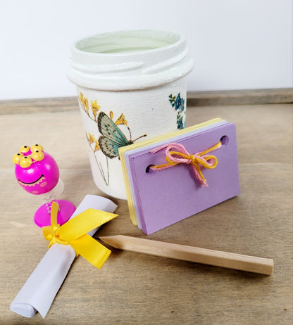 white painted jar with butterfly detail, lilac and yellow paper scrapbook, brown pencil, pink toy and a white rolled up note tied with yellow ribbon