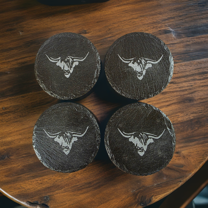 Four black slate coasters with cows head engraving on a wooden surface