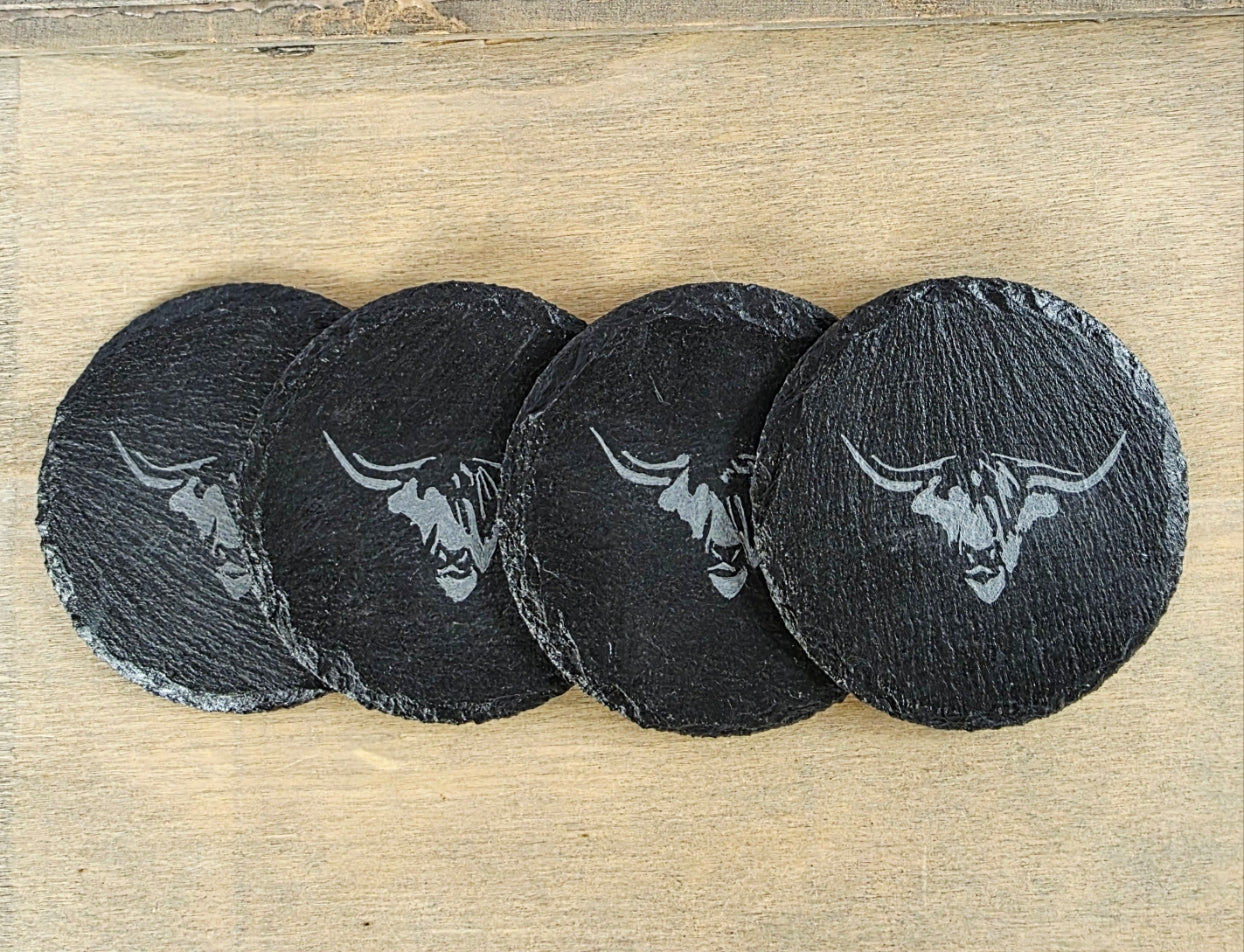 Four black round slate coasters with a cows head design on a wooden surface