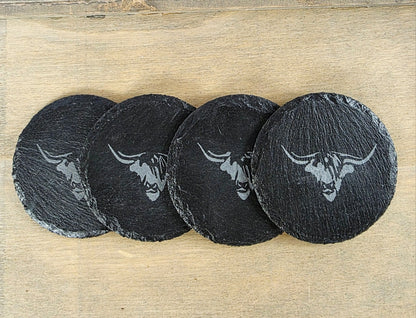 Four black round slate coasters with a cows head design on a wooden surface