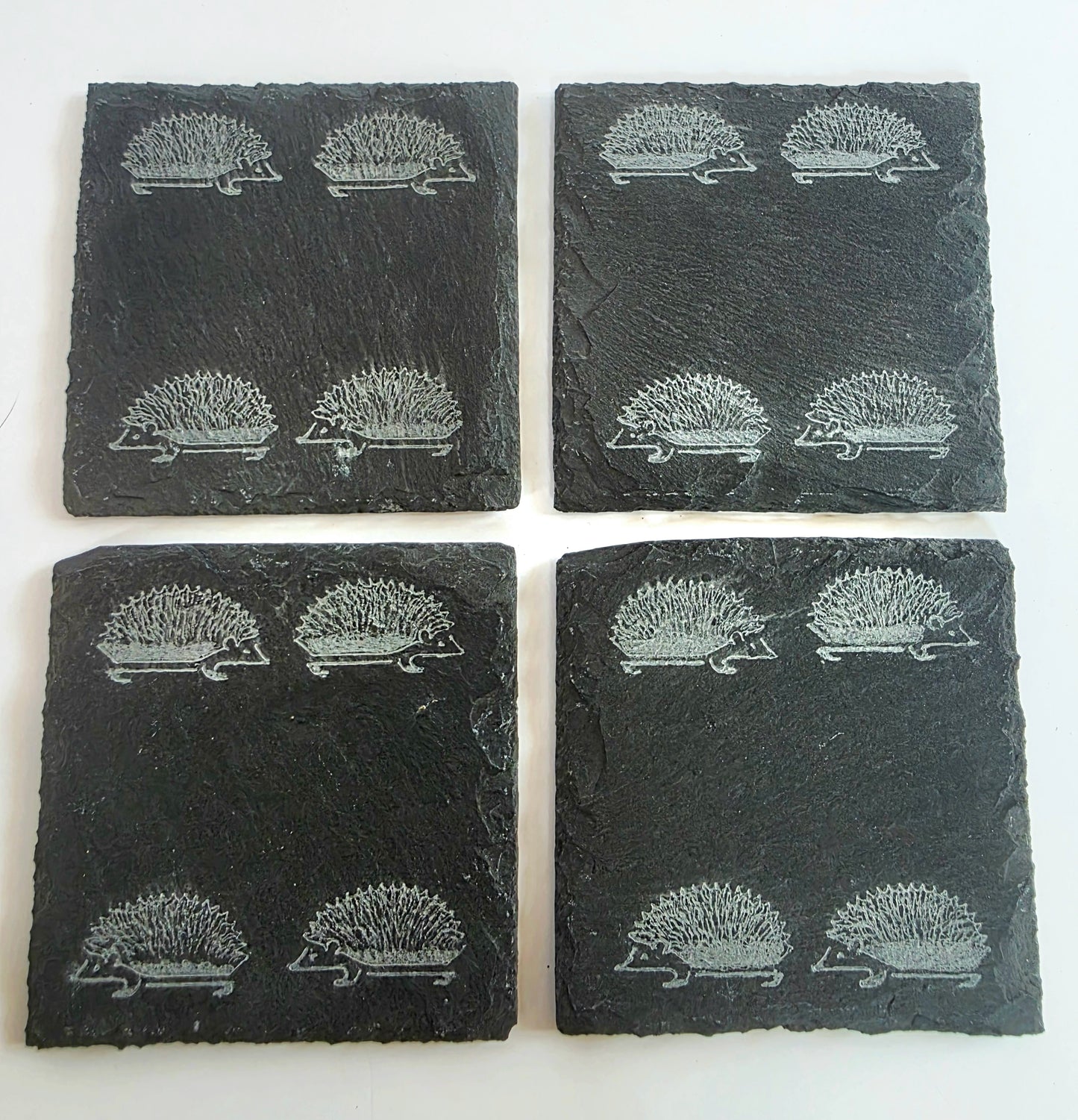 Four square dark slate coasters arranged in a 2x2 grid on a white background, each printed with a repeating hedgehog motif.