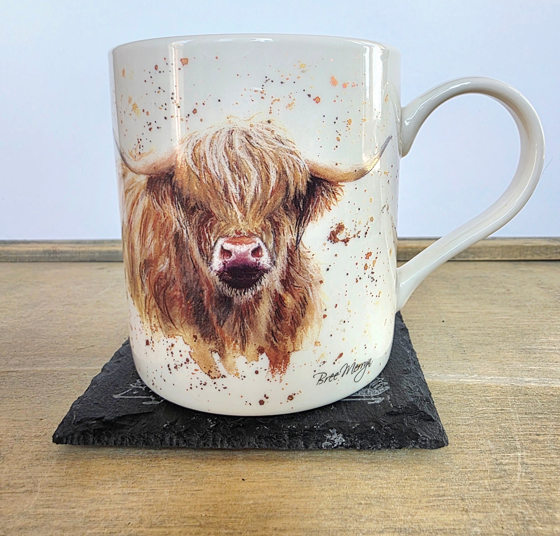 A ceramic mug featuring a detailed illustration of a Highland cow, displayed on a black slate coaster on a wooden table against a light blue background.