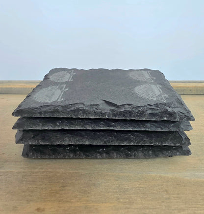 A stack of four dark gray slate coasters with natural rough edges and light gray engraved hedgehog motifs, displayed on a light wood surface against a pale blue background.