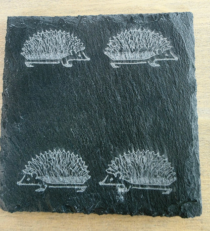 A square black slate coaster featuring an engraved pattern of four hedgehogs, shown on a light wood background.