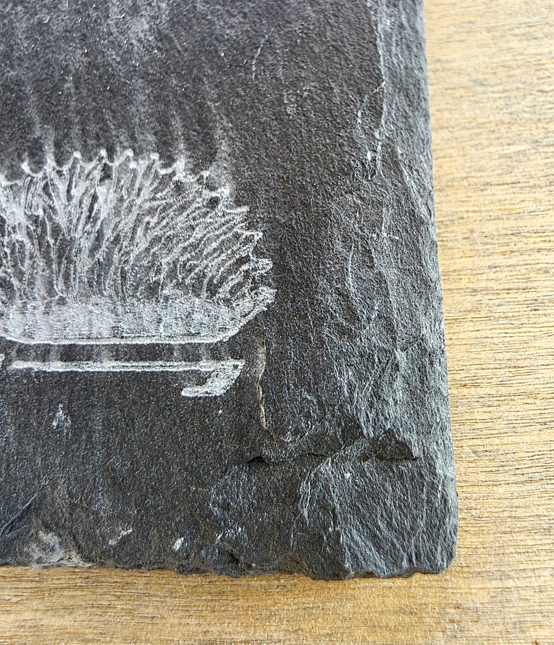 A close-up of a dark grey slate coaster with a white printed illustration of a cocktail glass and umbrella, resting on a light wood surface.