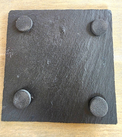 A square dark charcoal slate board with square corners and four circular textured rubber feet, shown on a light wood surface.