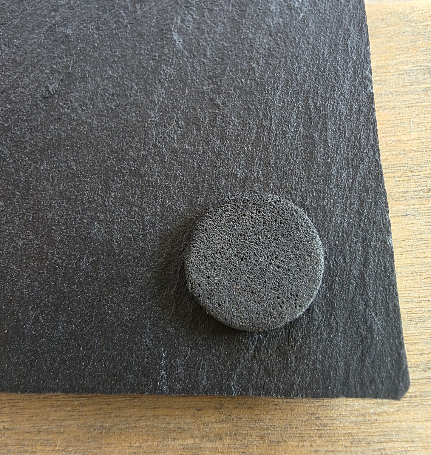 A close-up of a dark gray textured material sample with a circular foam-like pad placed on top, photographed on a light wood surface.