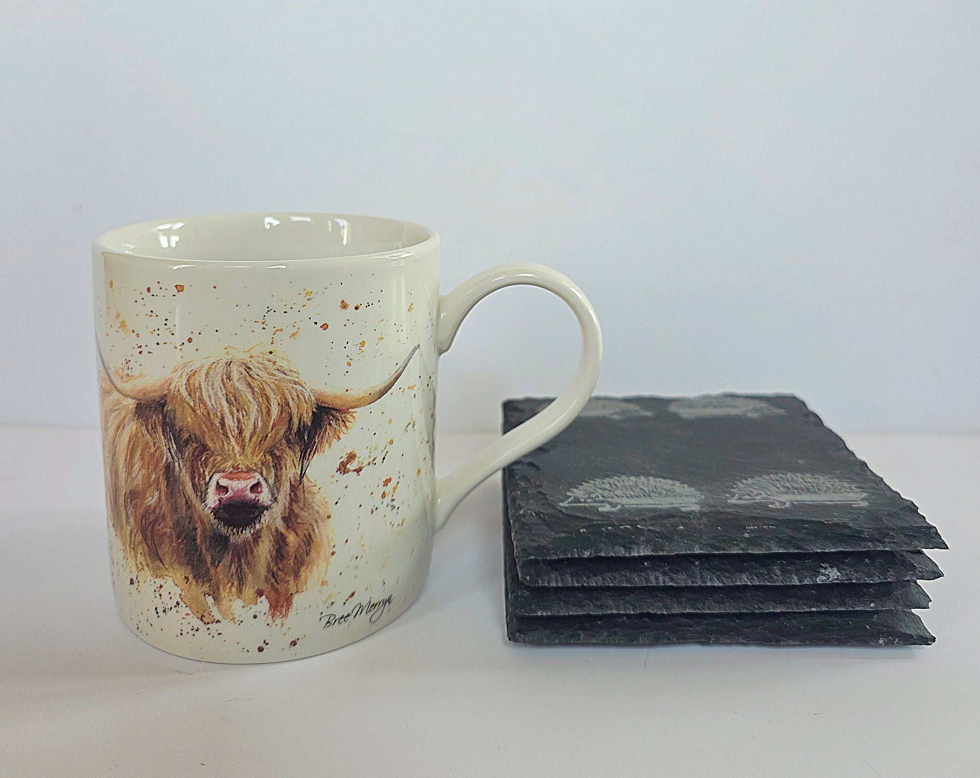 A ceramic mug with a painted Highland cow design sits next to a stack of dark slate coasters on a light gray background.