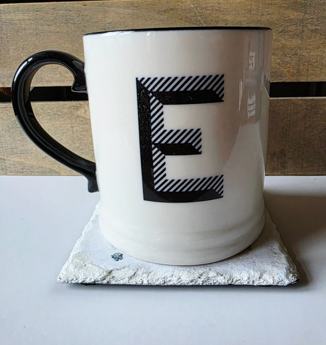 White mug with black handle and letter 'E' on a white surface with wooden background sitting on a white painted square slate coaster