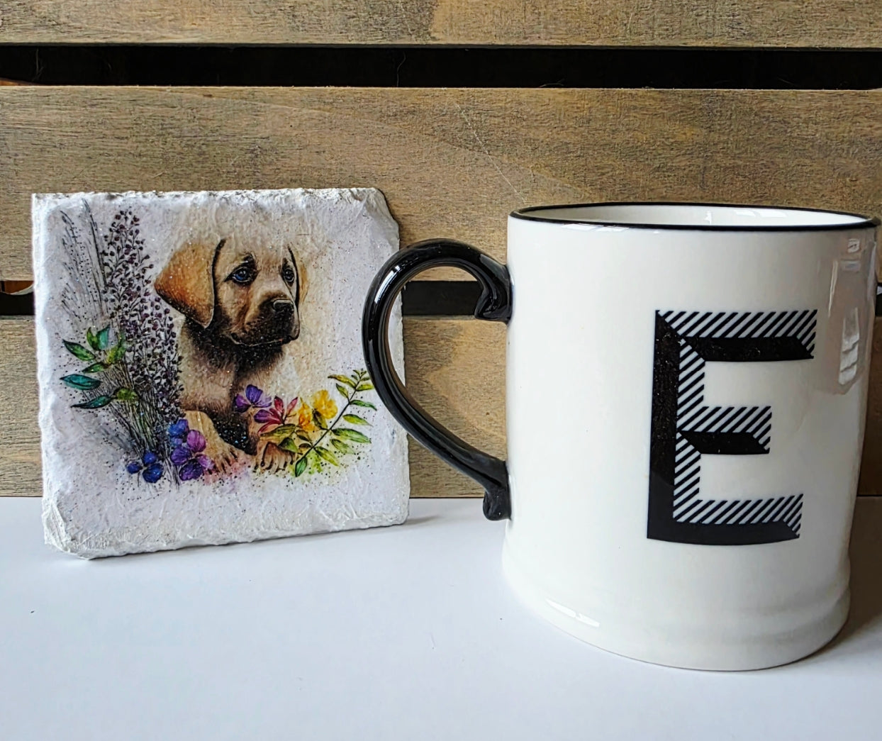 Mug with letter 'E' next to a white slate coaster with golden labrador puppy illustration next to a wooden surface