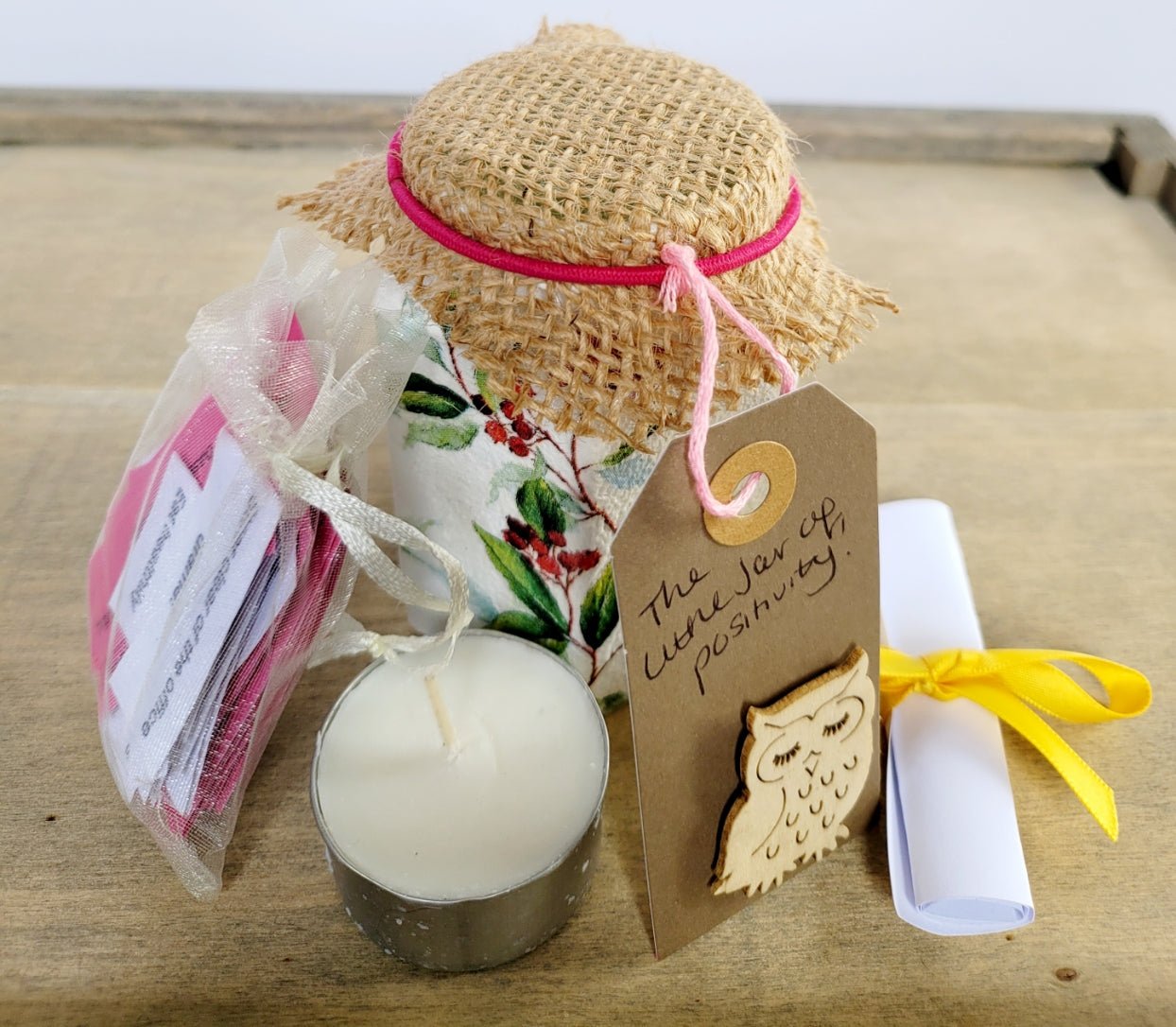 Small white painted jar with red and green berry leaf design. Pale organza bag with white and pink quotes, tealight candle, brown tag with 'little jar of positivity written on, white piece of rolled up paper, tied with yellow ribbon