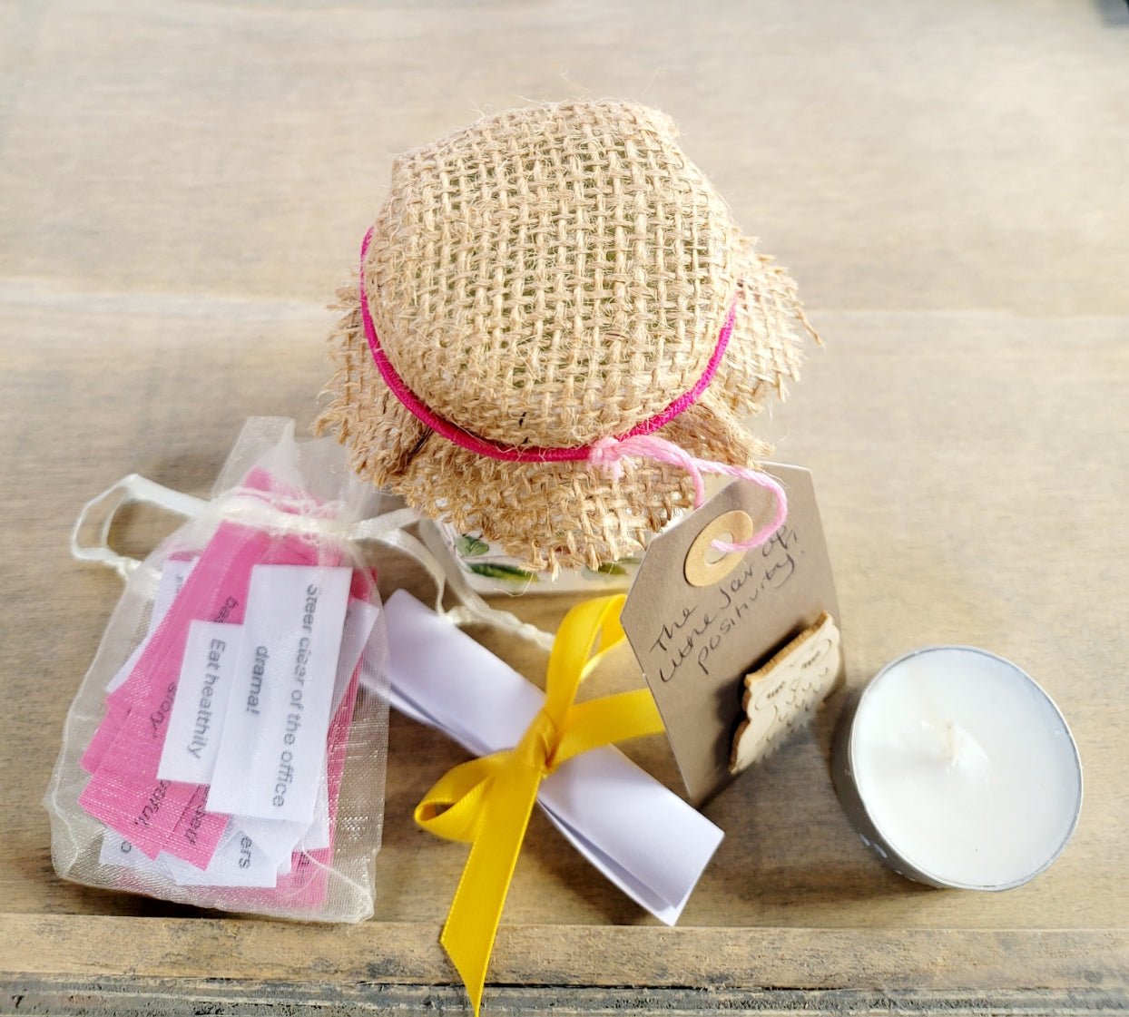 Hessian lid topped jar showing contents outside; pale organza bag with white/pink quotes, a white rolled up piece of paper tied with yellow ribbon and a white tealight candle.