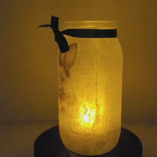 Rotating example of a Lit up glass jar with Hares in the snow detail, flameless candle and green bow