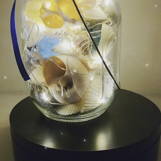 Jar filled with shells, pebbles and coloured glass stones. Lit up with lid and carry handle. Blue decorative ribbon