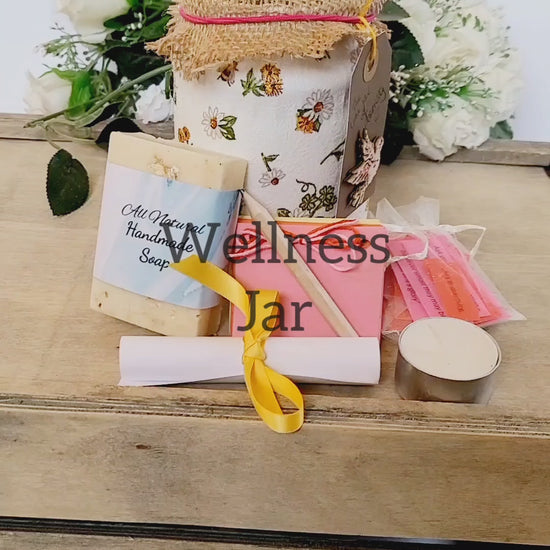 Vdeo of a White jar with bee and flower design, hessian lid with card label with a dove on it. Contents shown outside of the jar, cream soap, small multicoloured scrapbook, short wooden pencil, tealight candle, small organza bag containing coloured paper quotes, white rolled up instruction leaflet tied with yellow ribbon.